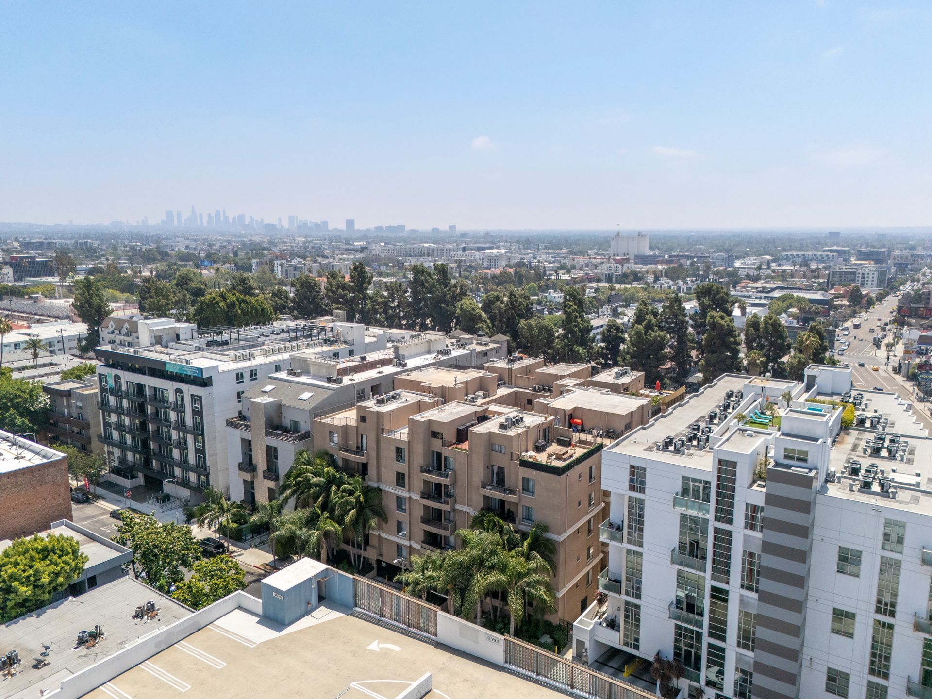 Aerial view of apartment buildings and palm trees in a sunny city neighborhood, with a distant skyline.