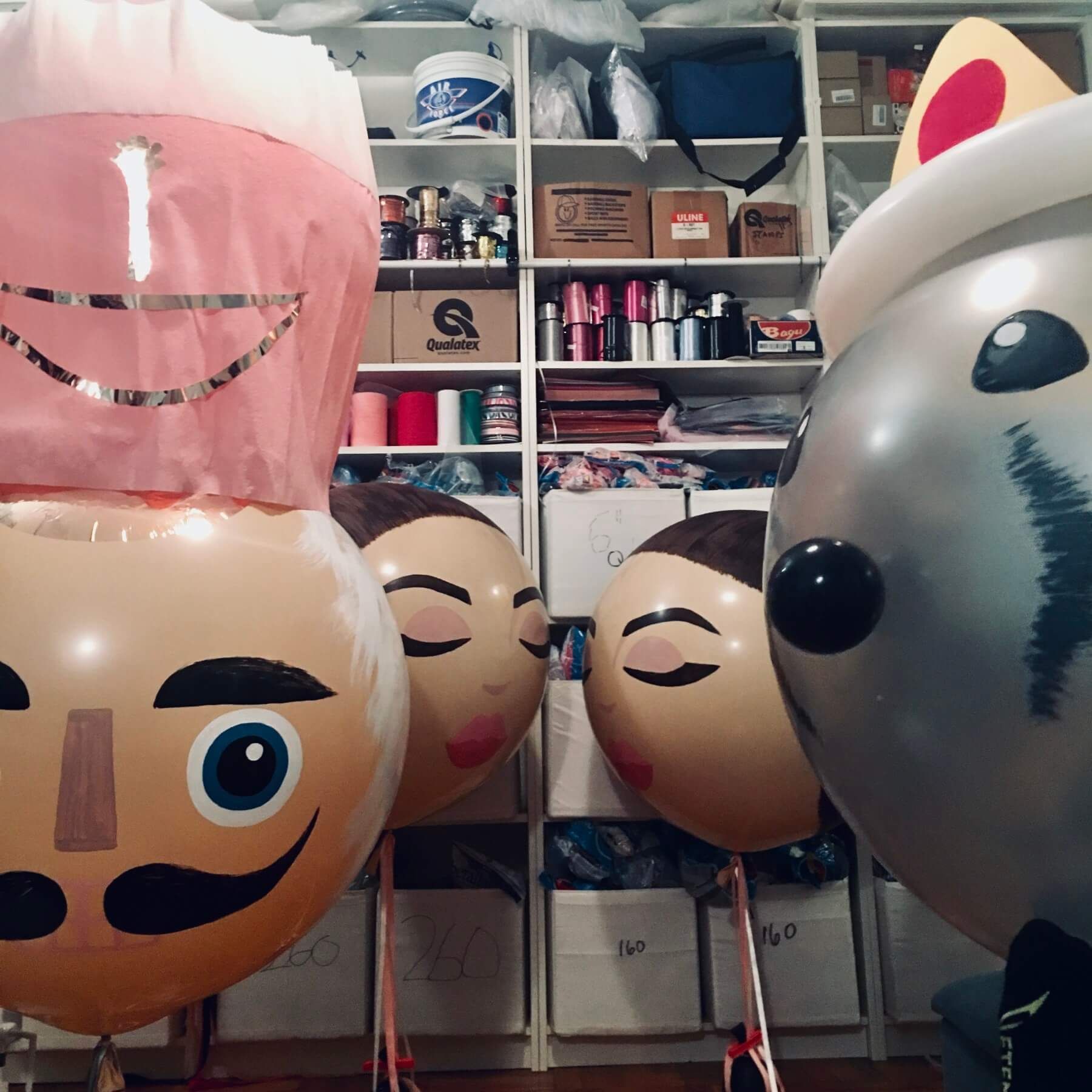 Balloons painted as people with closed eyes and a chef hat against a cluttered shelf backdrop.