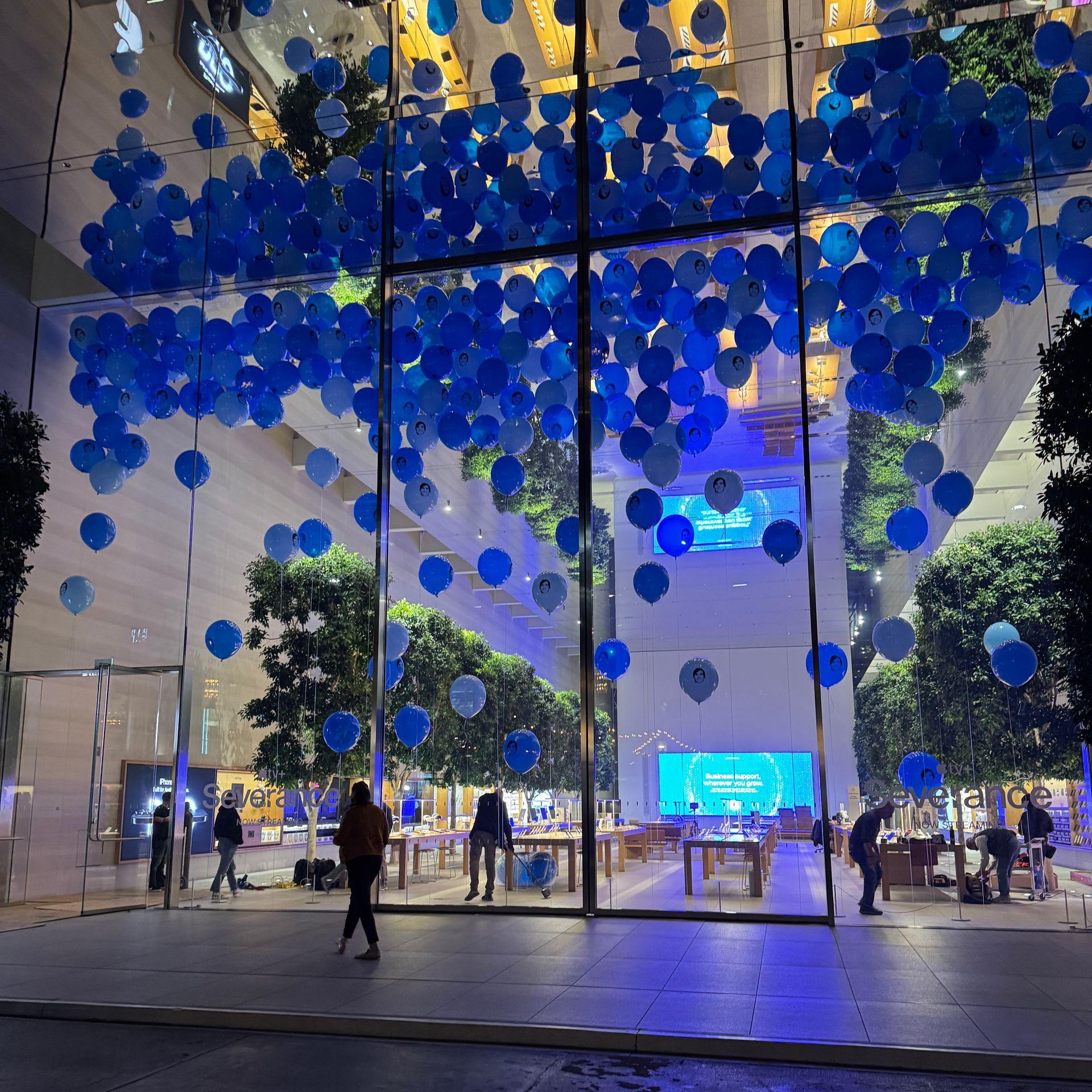 Apple store interior with blue balloons, people browsing, and trees.