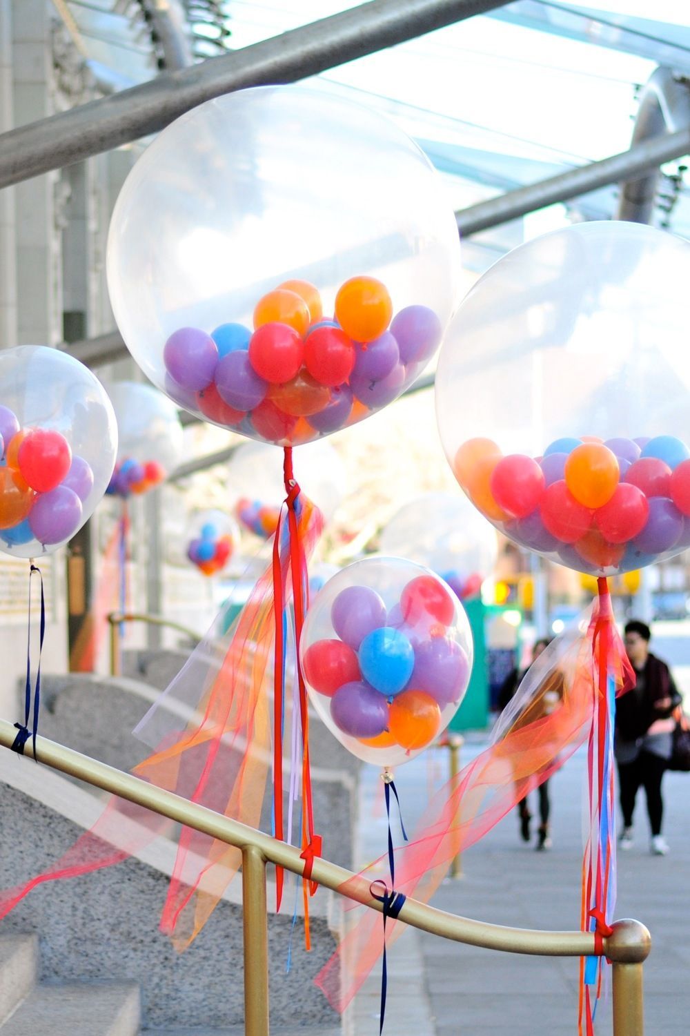 Clear balloons filled with colorful smaller balloons, hanging from a railing, outdoors.