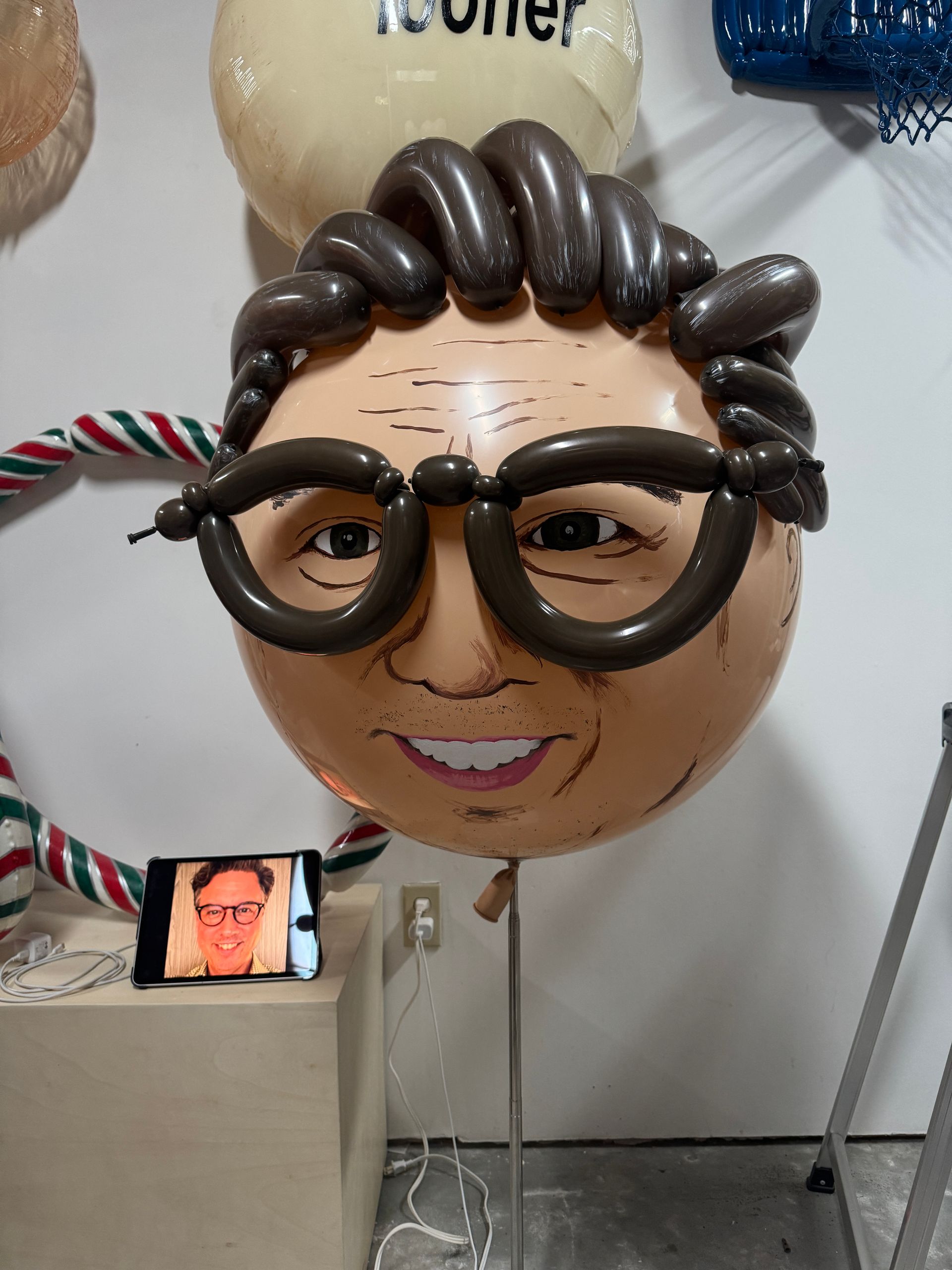 A balloon sculpture of a man's face with brown hair, glasses, and a smiling expression. A phone displays the man's face.