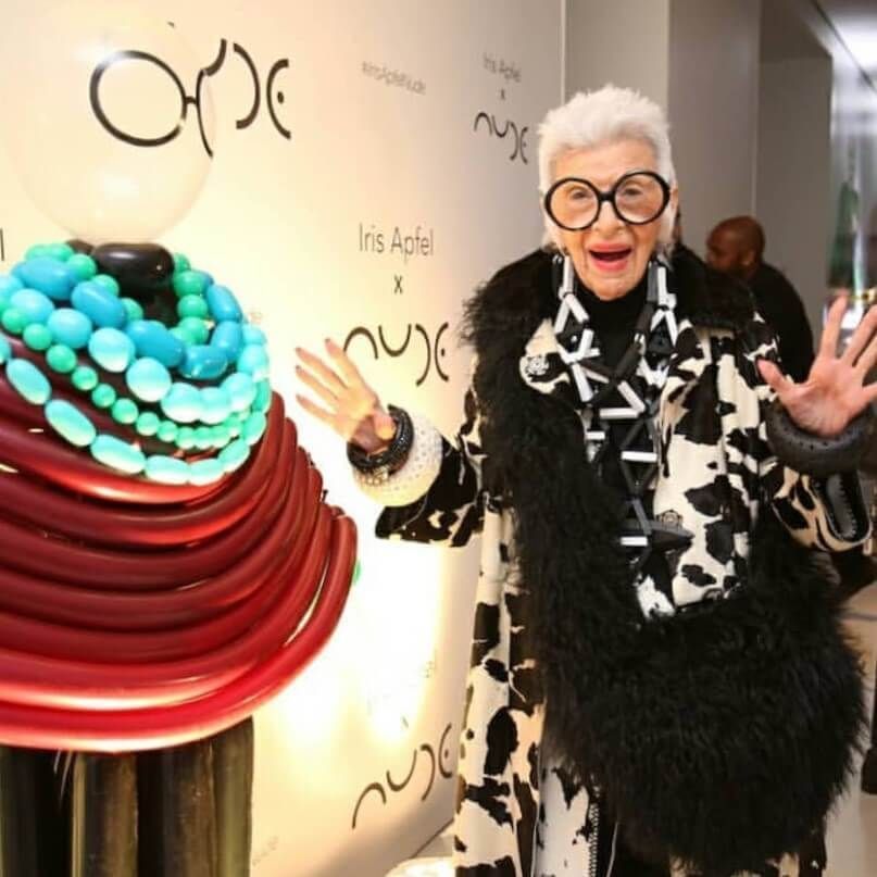 A woman wearing glasses and a fur coat stands in front of a wall that says 