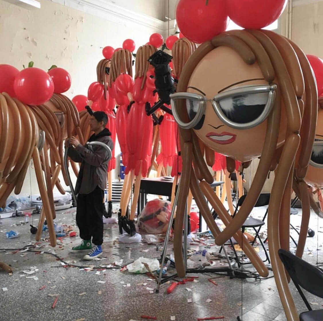 Man adjusts a large balloon sculpture of a woman with sunglasses and tomato balloons on top of the head in a studio.