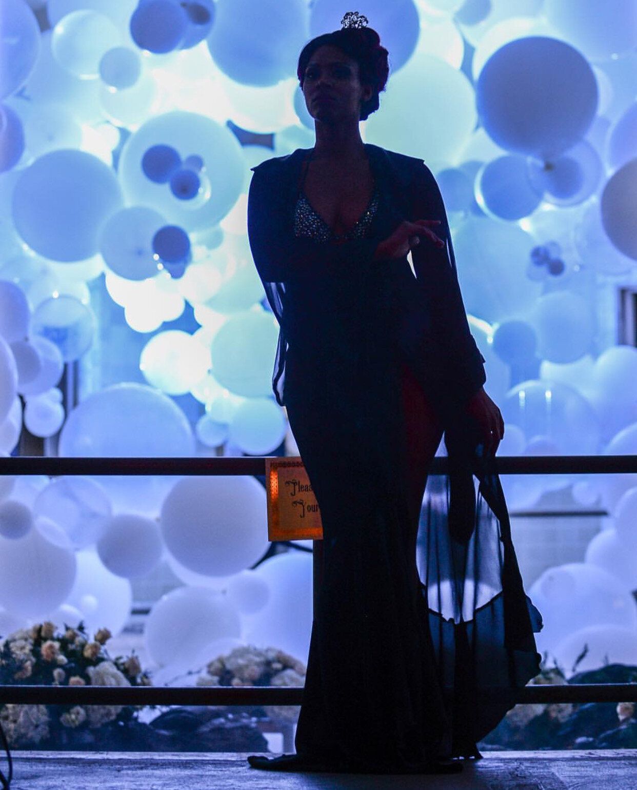 Woman in formal wear stands near balloons; a red shawl is draped over her arm.