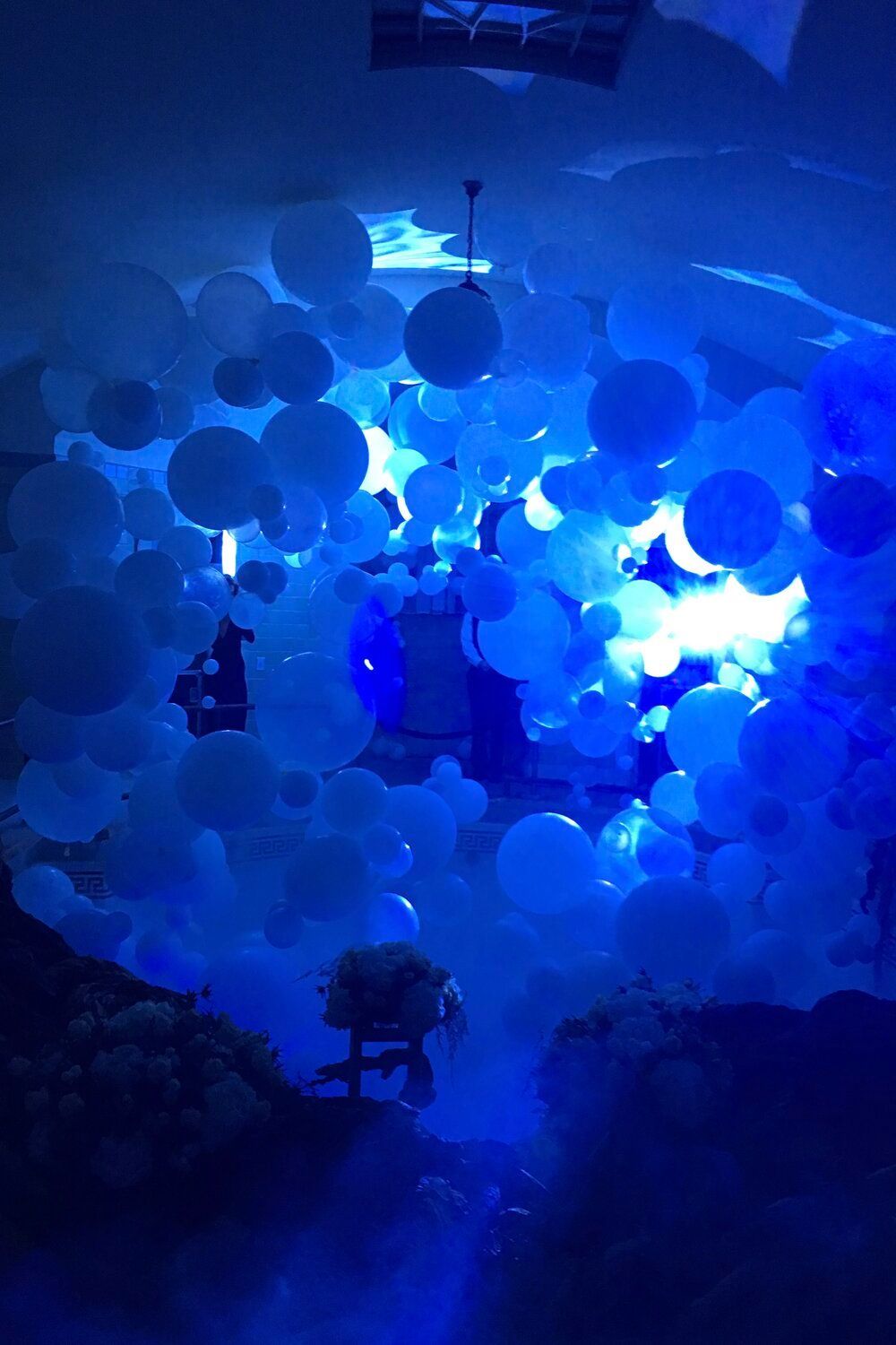 Blue lit room filled with numerous white and blue balloons; a person stands inside.