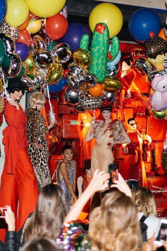 Models on a makeshift stage, with audience members. Balloons and various decorations fill the space.