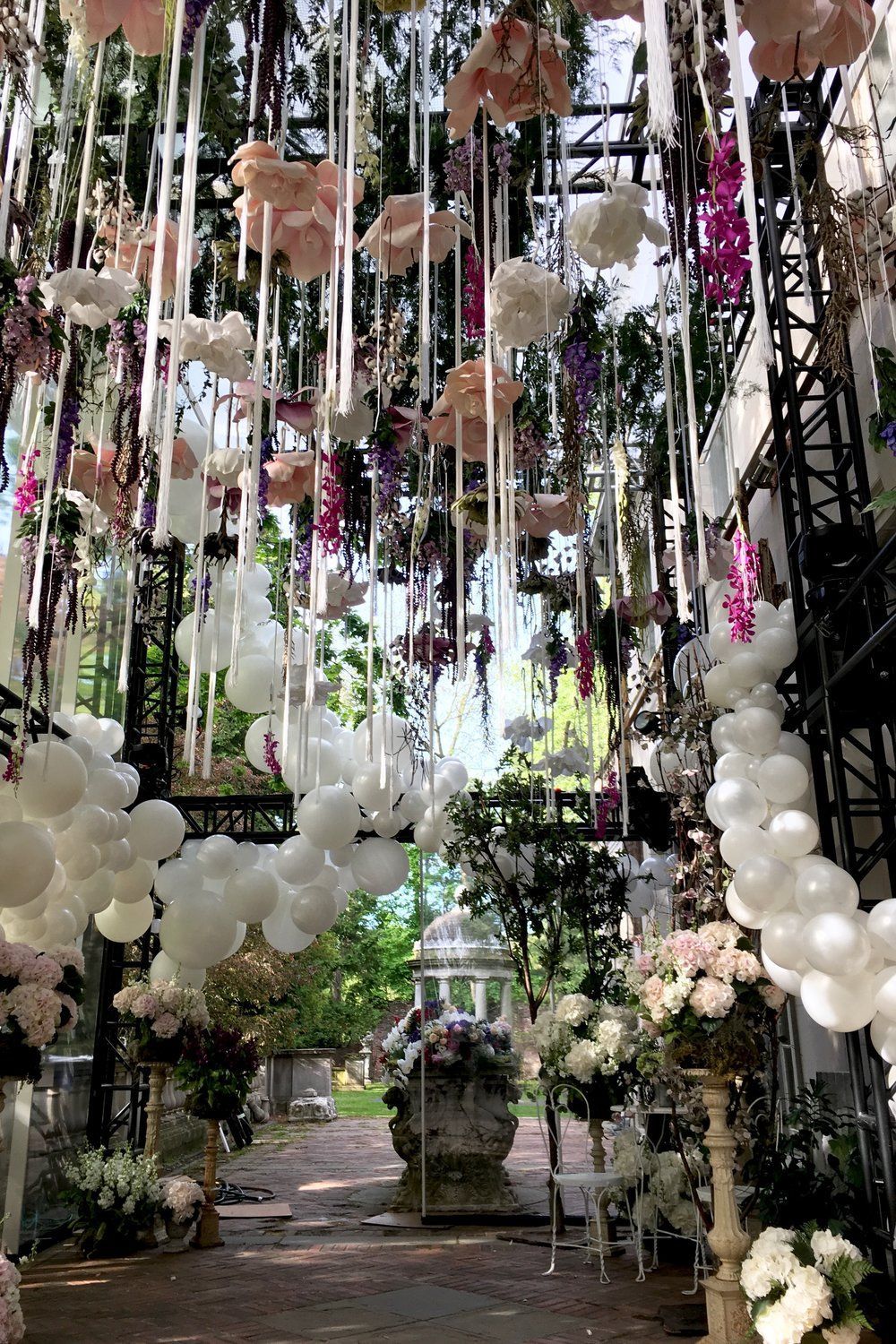 An outdoor floral archway decorated with white balloons, ribbons, and pink and purple flowers.