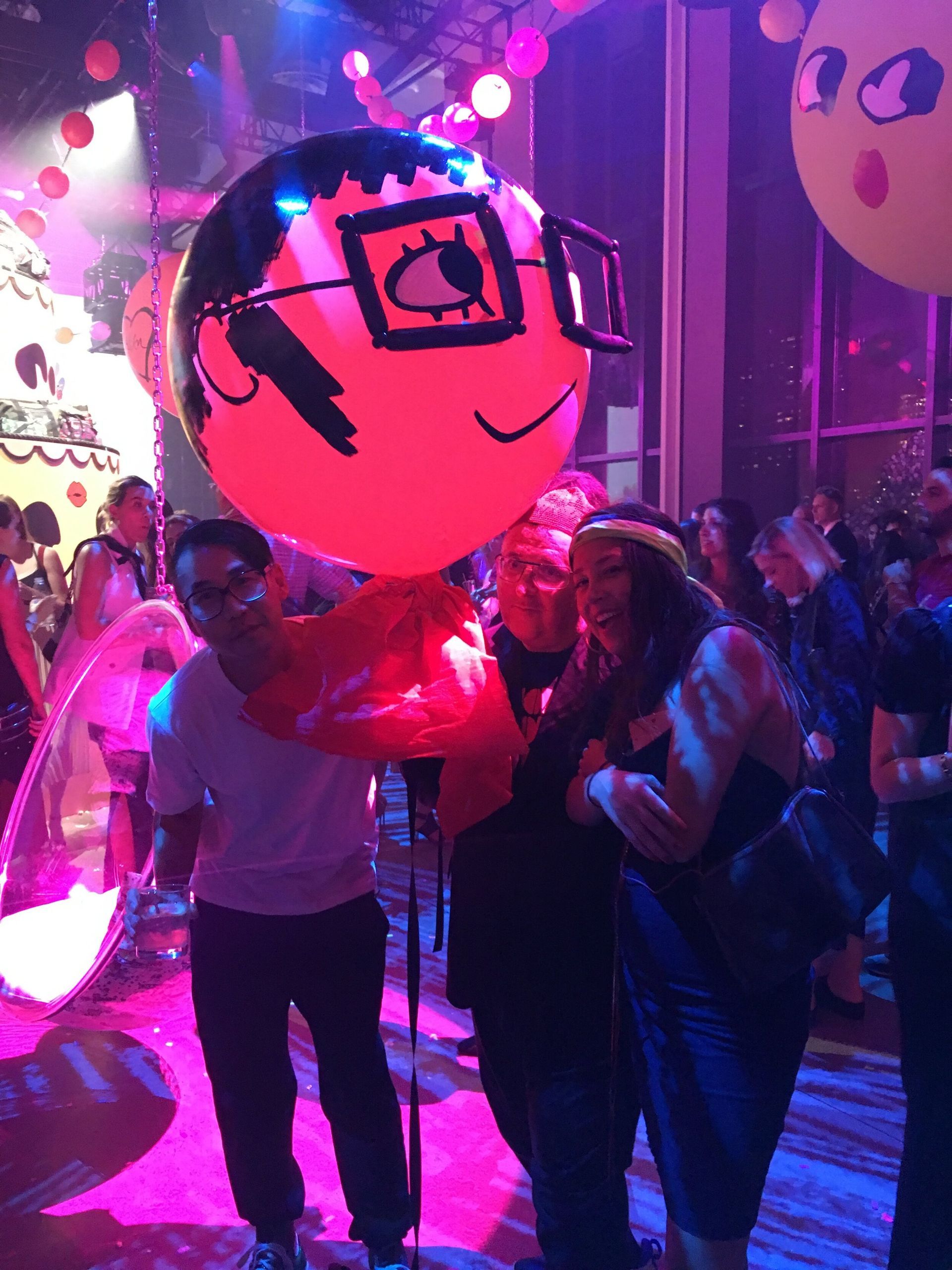 People holding up a large, round red balloon with a cartoon face at an event with lights and decorations.