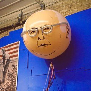 Round sculpture of a man's face wearing glasses, drawn on a sphere, mounted on a blue wall.