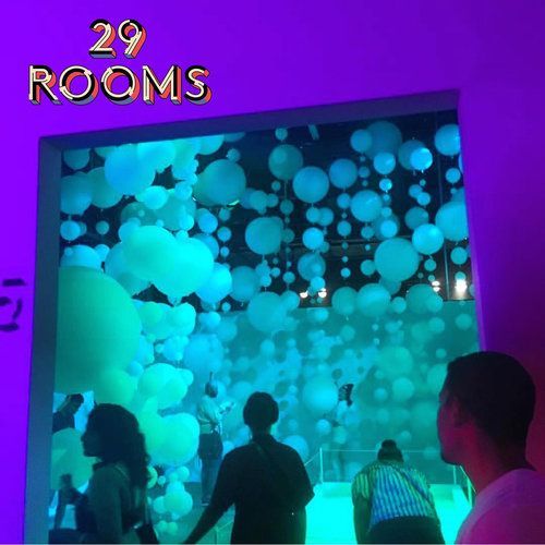 People in a room with blue-green spheres filling a window. Purple and green lighting. 29 Rooms text.