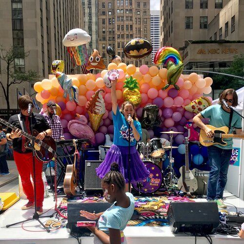 A band performs on an outdoor stage decorated with balloons. A woman sings and a girl dances in front of them.