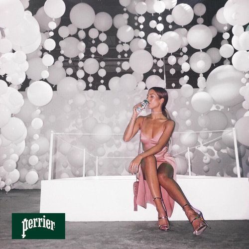 Woman sips water, seated on a white bench, surrounded by white balloons. The Perrier logo is in the bottom-left corner.