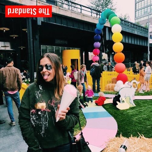 Woman with a sweet treat at an outdoor event with a rainbow arch, holding her tongue out.