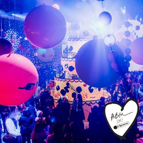 Party with large balloons, cake, and crowd in colorful lighting.