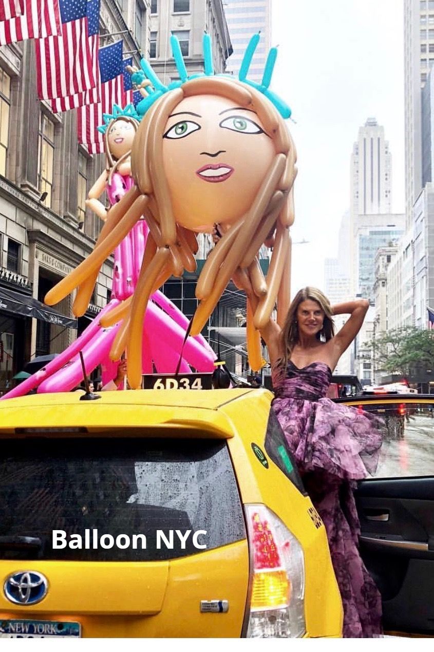 Woman in dress poses with balloon sculpture on yellow taxi in NYC.