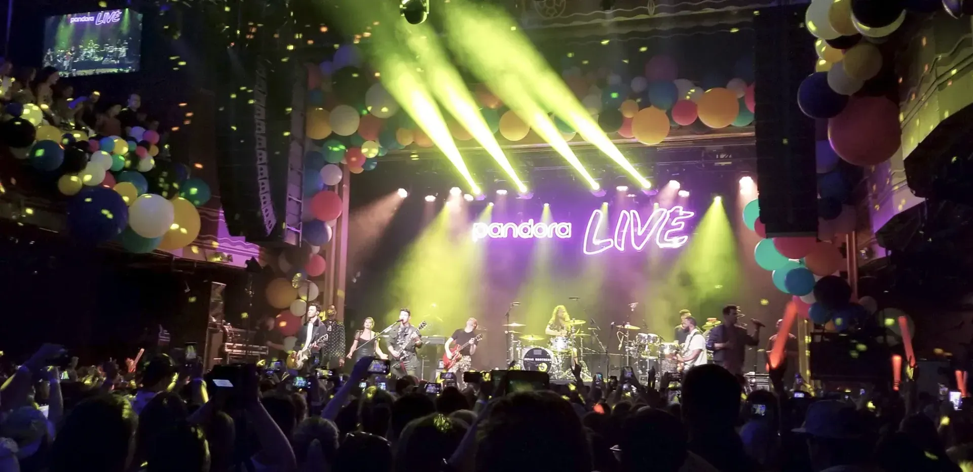 A band performing on stage at a concert. Stage lights, balloons, and a crowd.