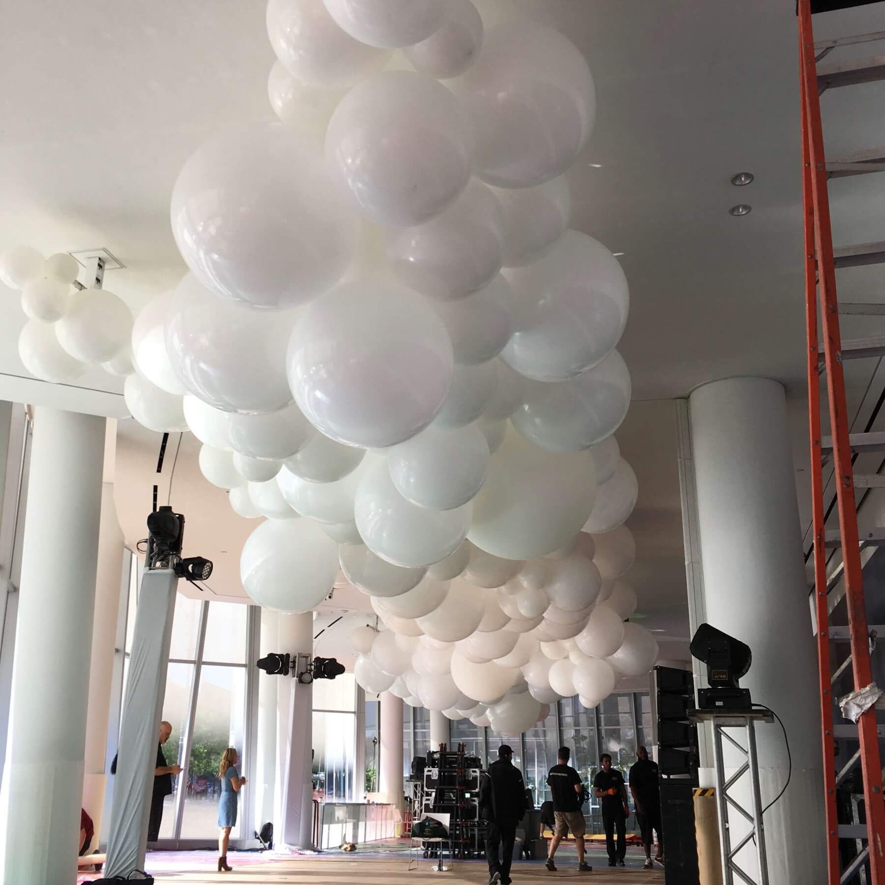 White balloons clustered on a ceiling; people in a room with pillars, stage equipment and windows.