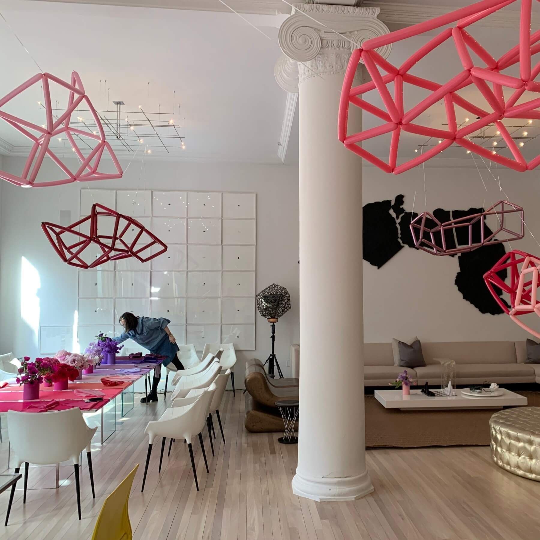 Room with pink geometric sculptures, a long table set for an event, and a person arranging flowers.