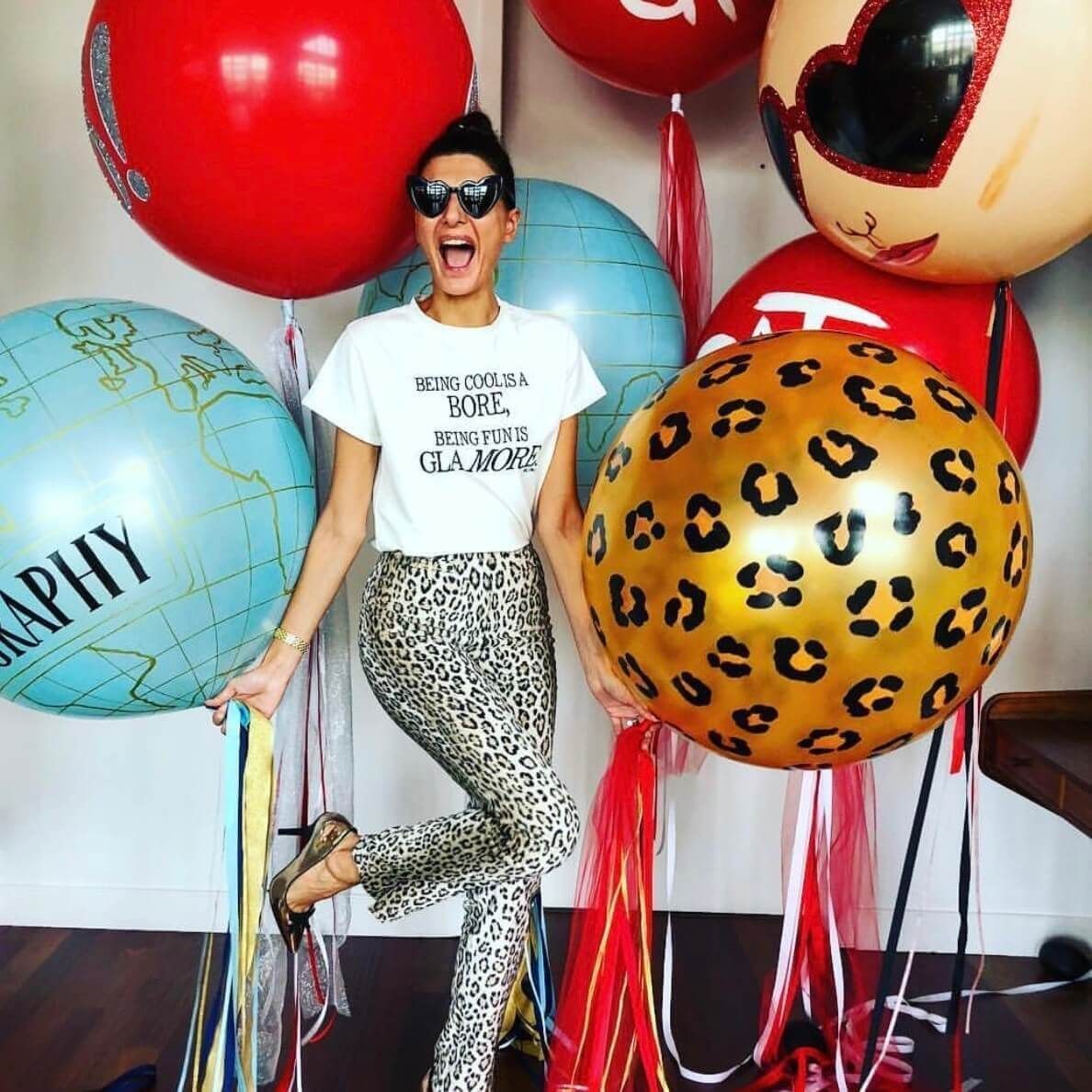 Woman in leopard pants smiles, surrounded by colorful balloons, some with text and patterns.