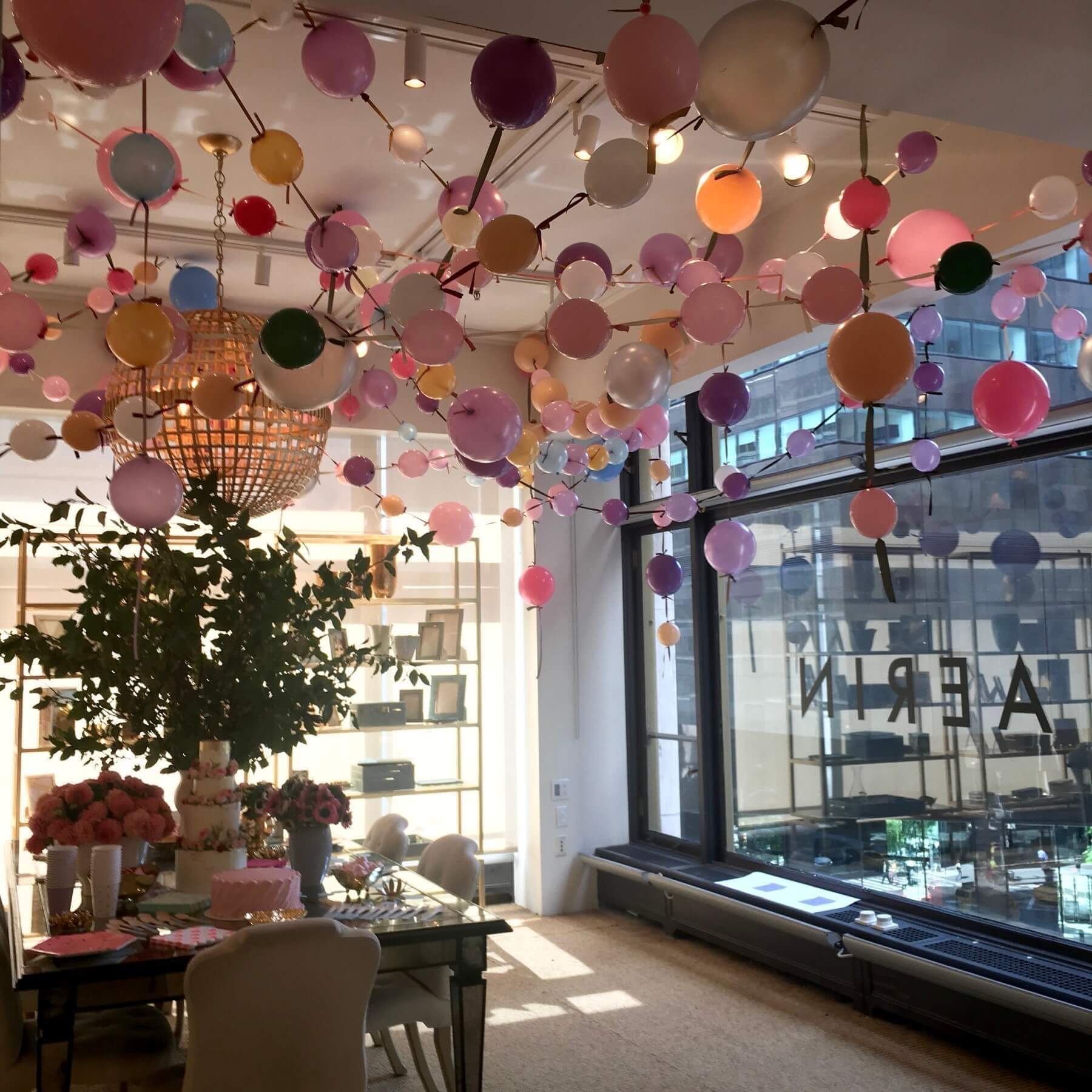 Room decorated with pastel balloons, chandelier, and a table set for a celebration. Sunlight streams through a large window.