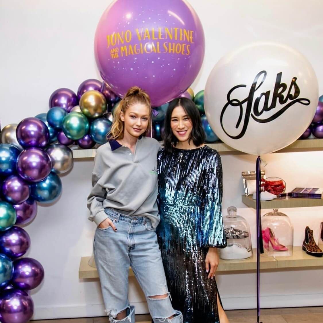 Two women pose at a shoe event; one in gray sweater and ripped jeans, the other in a sequined gown, surrounded by balloons.