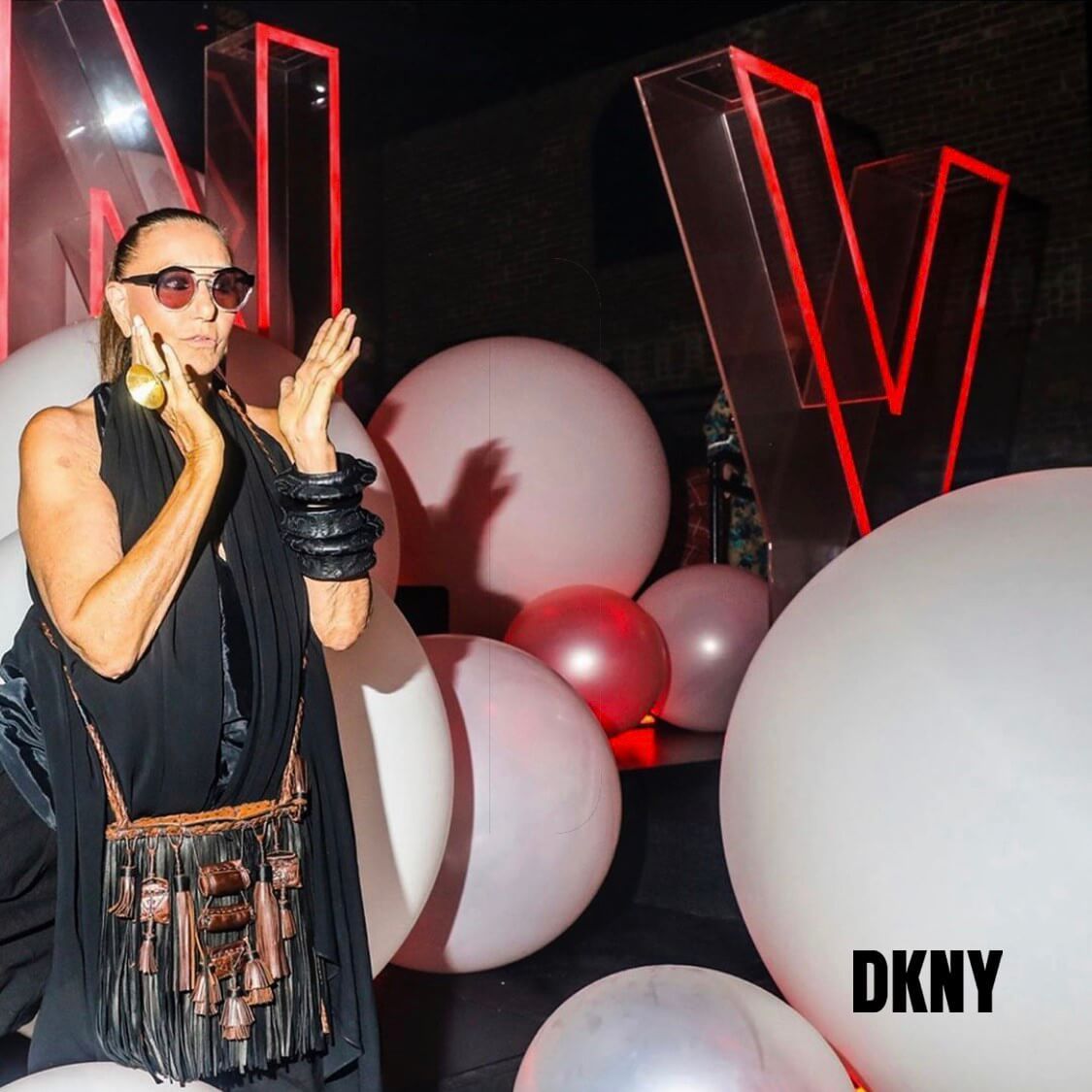 Woman in sunglasses and black outfit, gesturing in front of white and pink balloons and 