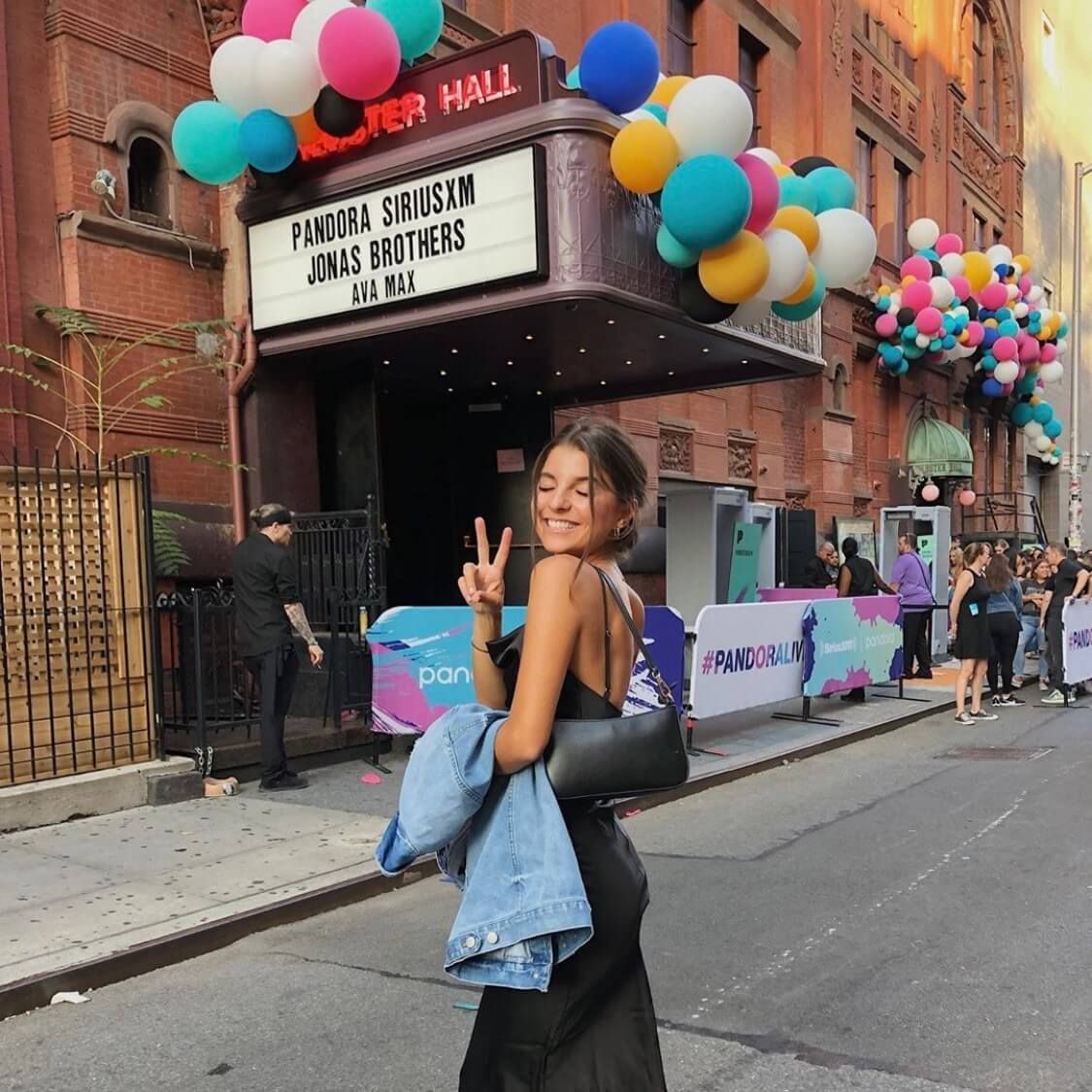 Woman in a black dress giving a peace sign, outside a theater decorated with balloons.