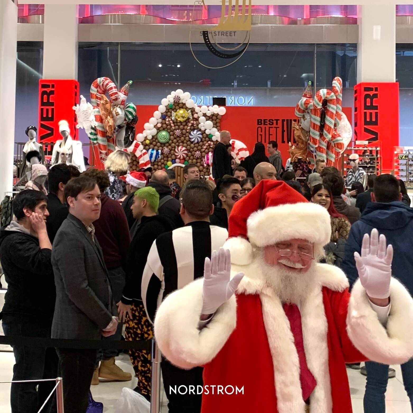 Santa Claus waving in a crowded Nordstrom store, surrounded by Christmas decorations and shoppers.
