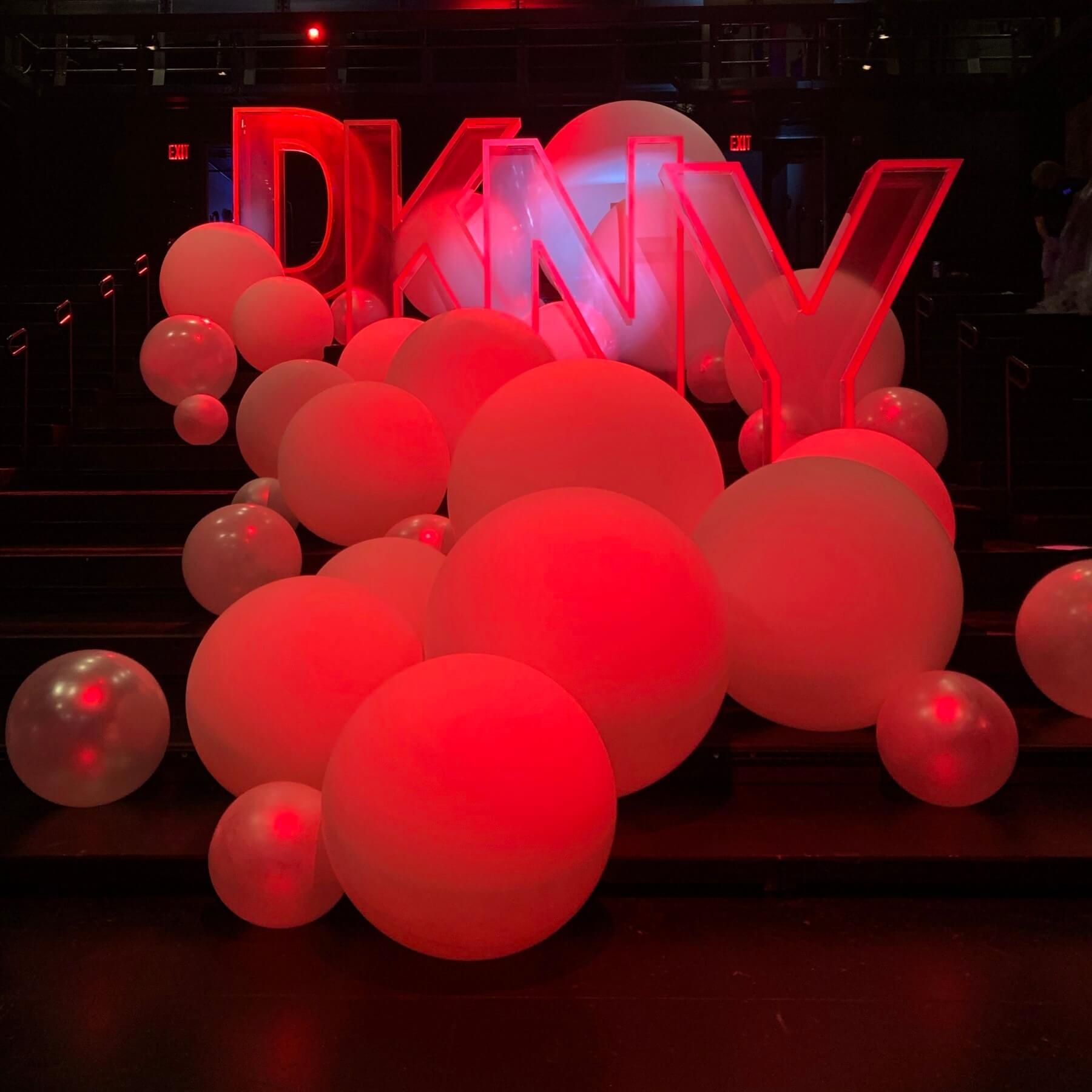 DKNY logo with red neon lights, surrounded by red and white balloons. Dark setting.