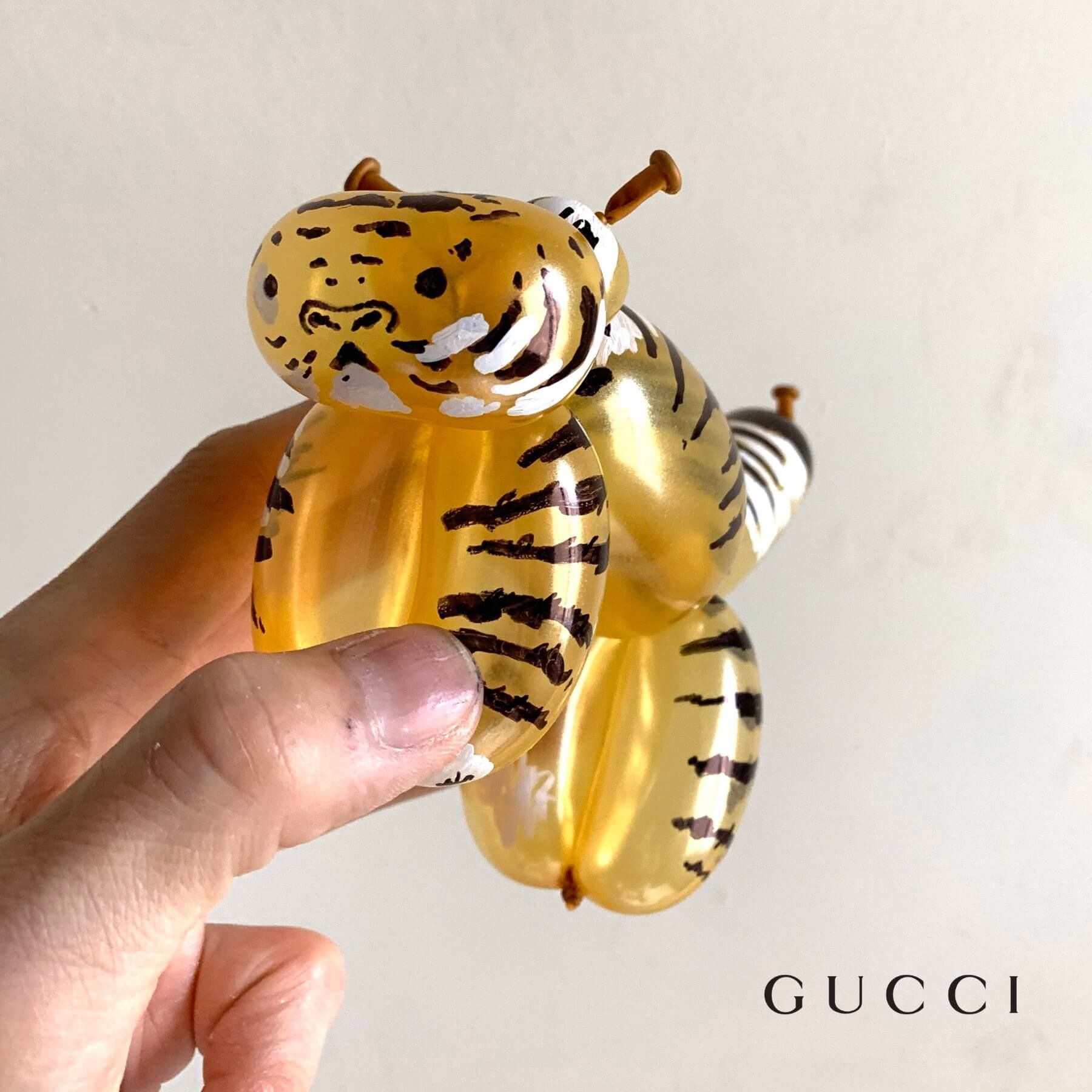 Balloon tiger, yellow and black stripes, held by a hand. Gucci logo visible.