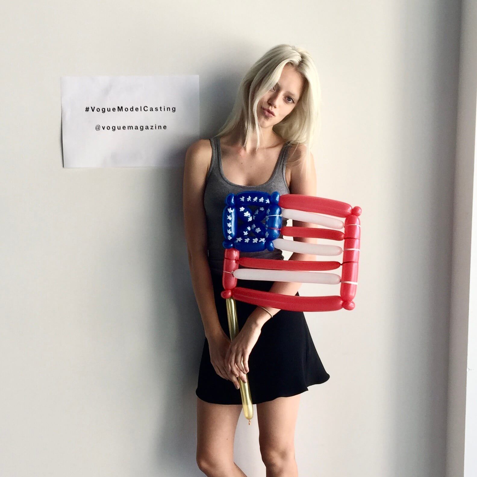 Blonde woman holding balloon American flag, wearing grey tank top and black skirt. White wall background.