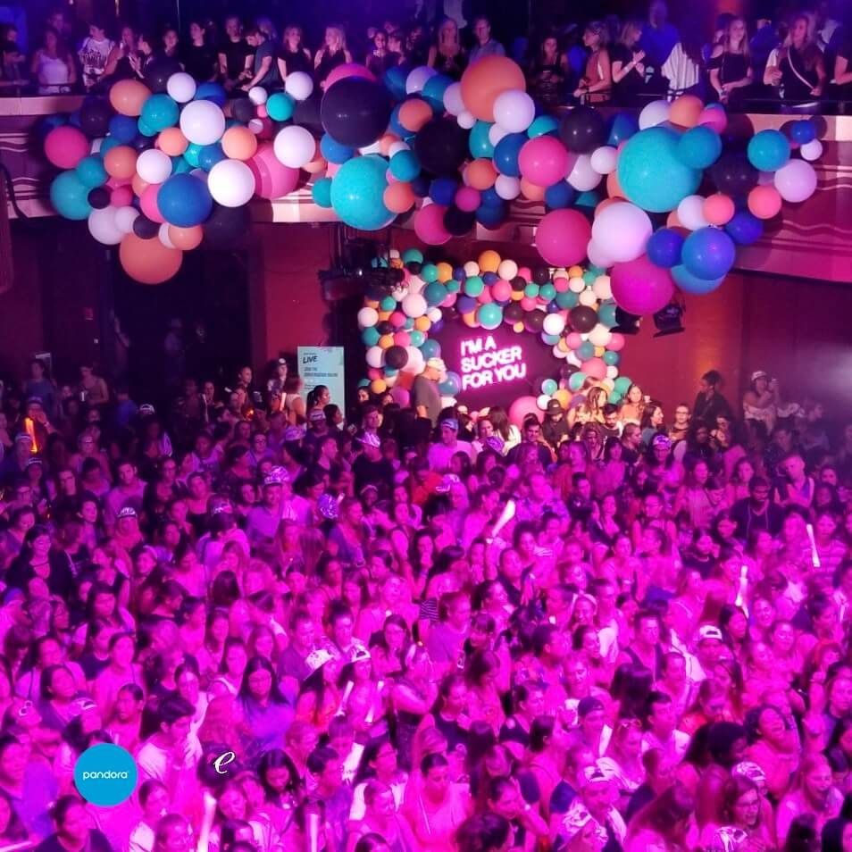 Large crowd at a venue; many people lit with pink light; balloons decorate the stage.