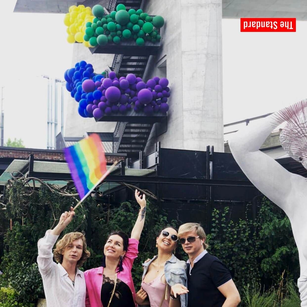 Four people waving; holding a rainbow flag, under a structure decorated with rainbow balloons.