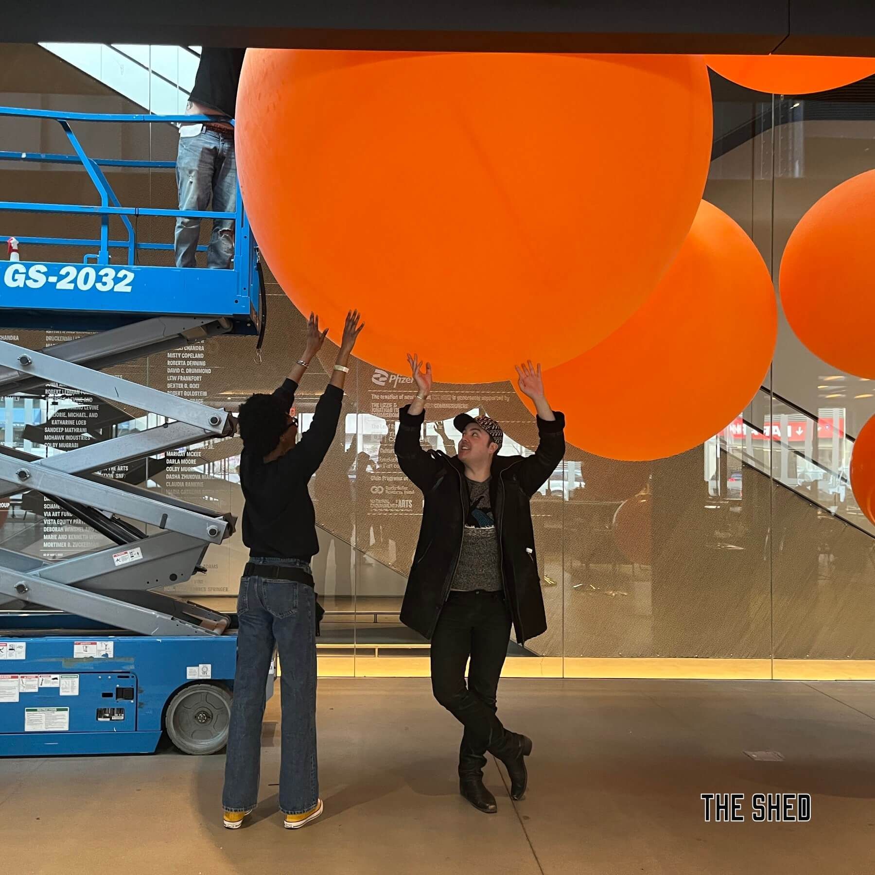 Two people install large orange spheres near a patterned wall, a scissor lift is visible in the background.