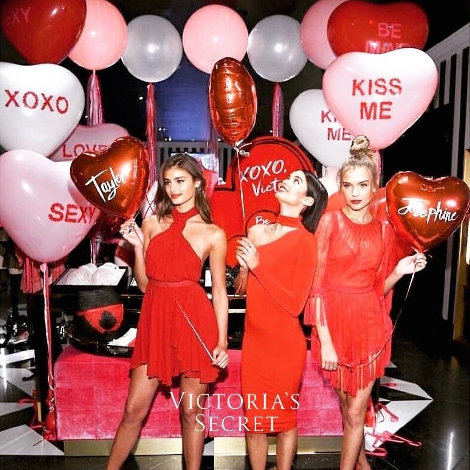 Three women in red dresses with Valentine's Day balloons in a Victoria's Secret setting.