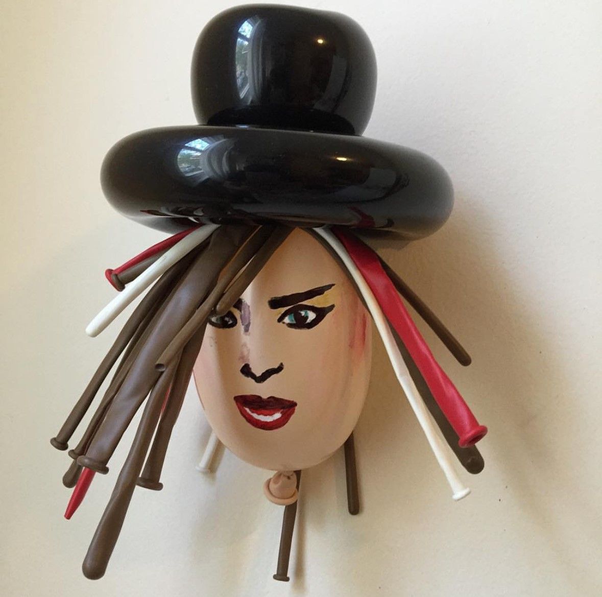 Sculpture of a face with a black hat, nail-like hair, and a neutral expression. Mounted on a white wall.