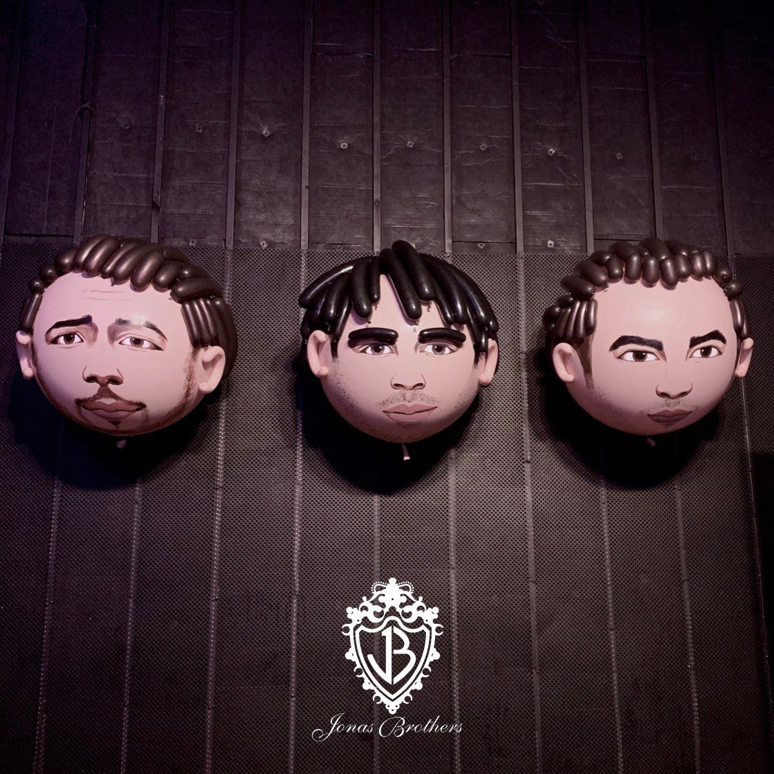 Three spherical faces mounted on a dark textured surface. The faces have distinct hairstyles and expressions. The bottom displays a logo.
