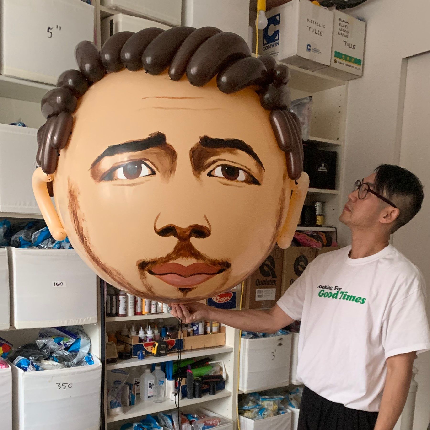 Man holding a large balloon head. The balloon is a tan face with painted features; the hair is made of brown balloons.