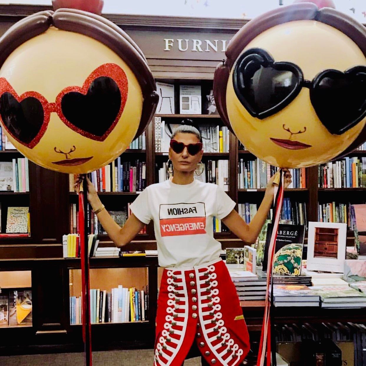 Woman holding two balloon heads with heart-shaped sunglasses in a bookstore. She wears a white tee and red pants.