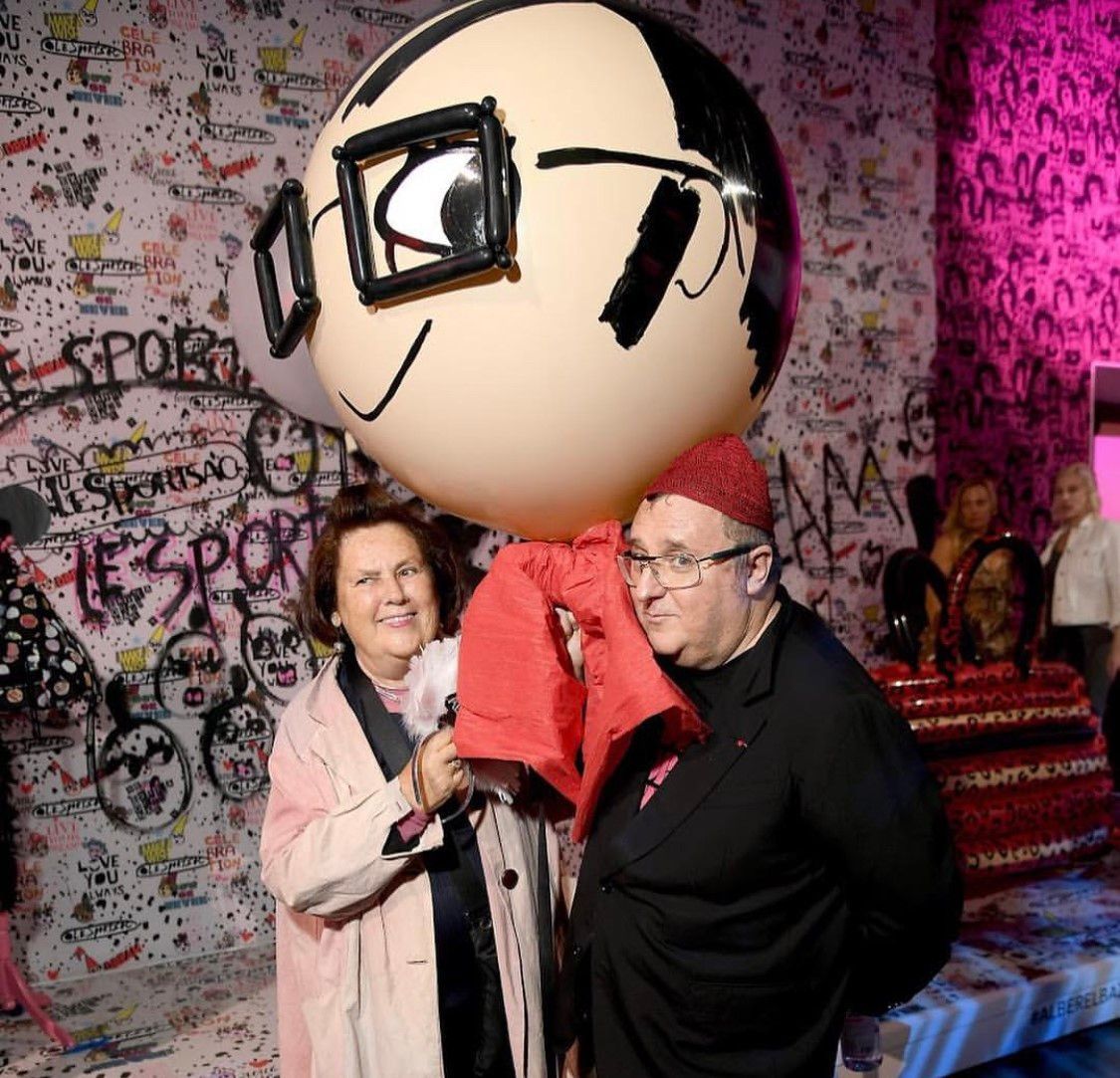 Two people pose with a large cartoon head balloon in a graffiti-covered room.
