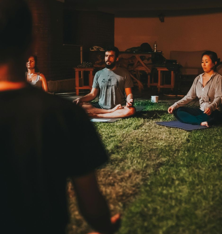 People meditating on mats outdoors at dusk, with warm lighting and a grassy setting