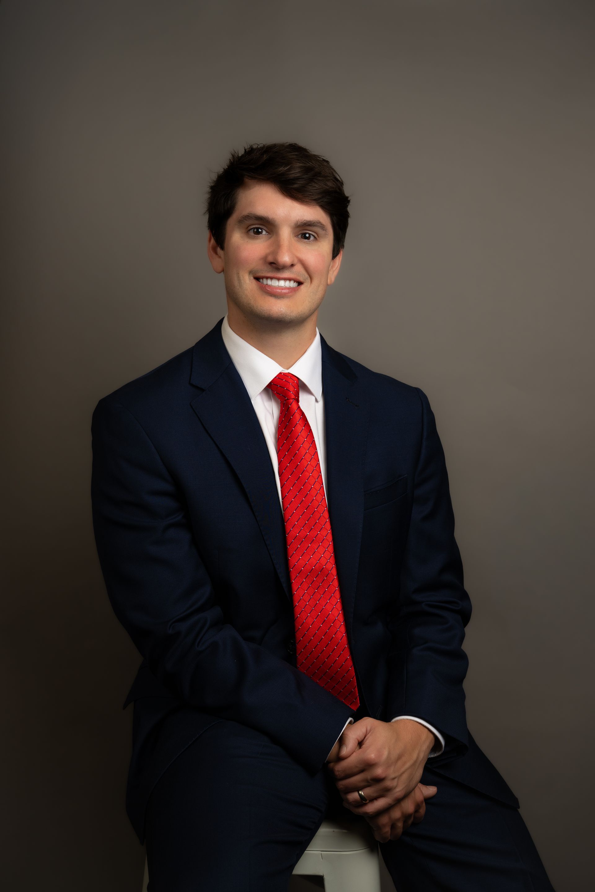 Man in a dark suit with a red tie, seated and smiling against a plain gray background