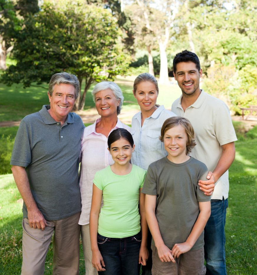 Family of seven smiling in a park, including two adults, two children, and grandparents, amidst green trees.
