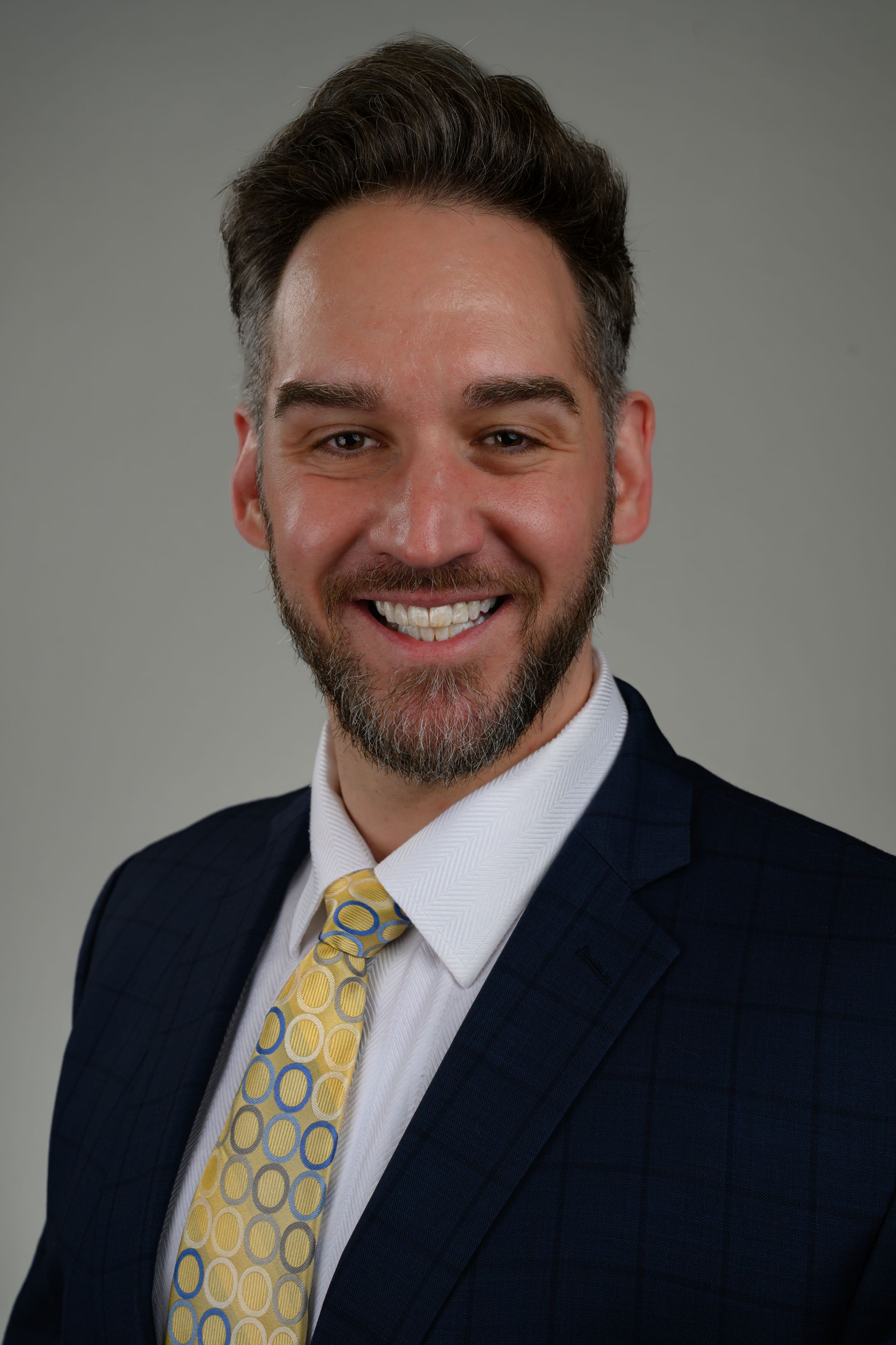 Man in navy suit, white shirt, and yellow patterned tie smiling at the camera.