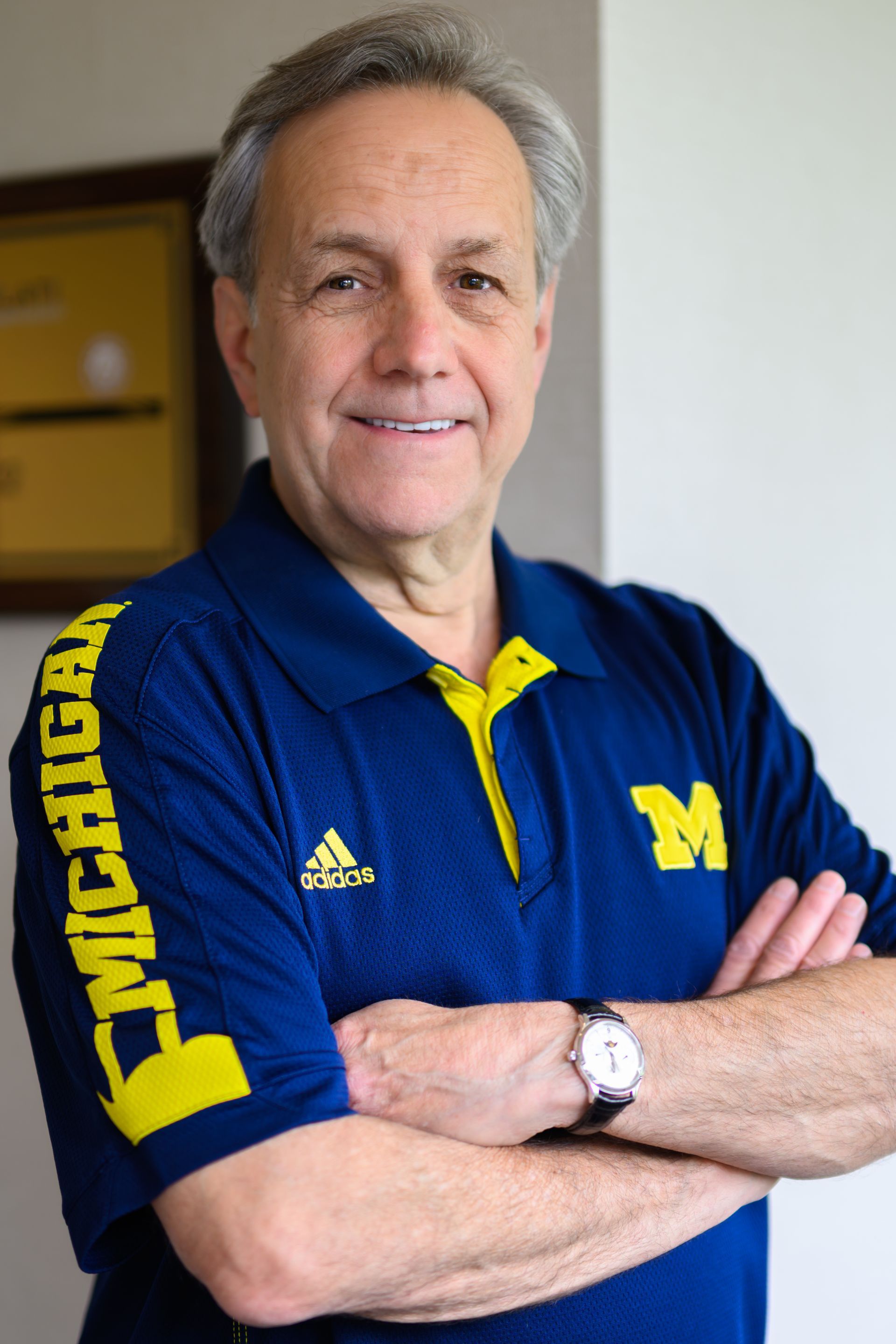 Man in blue and yellow Michigan shirt with arms crossed, smiling, indoors.