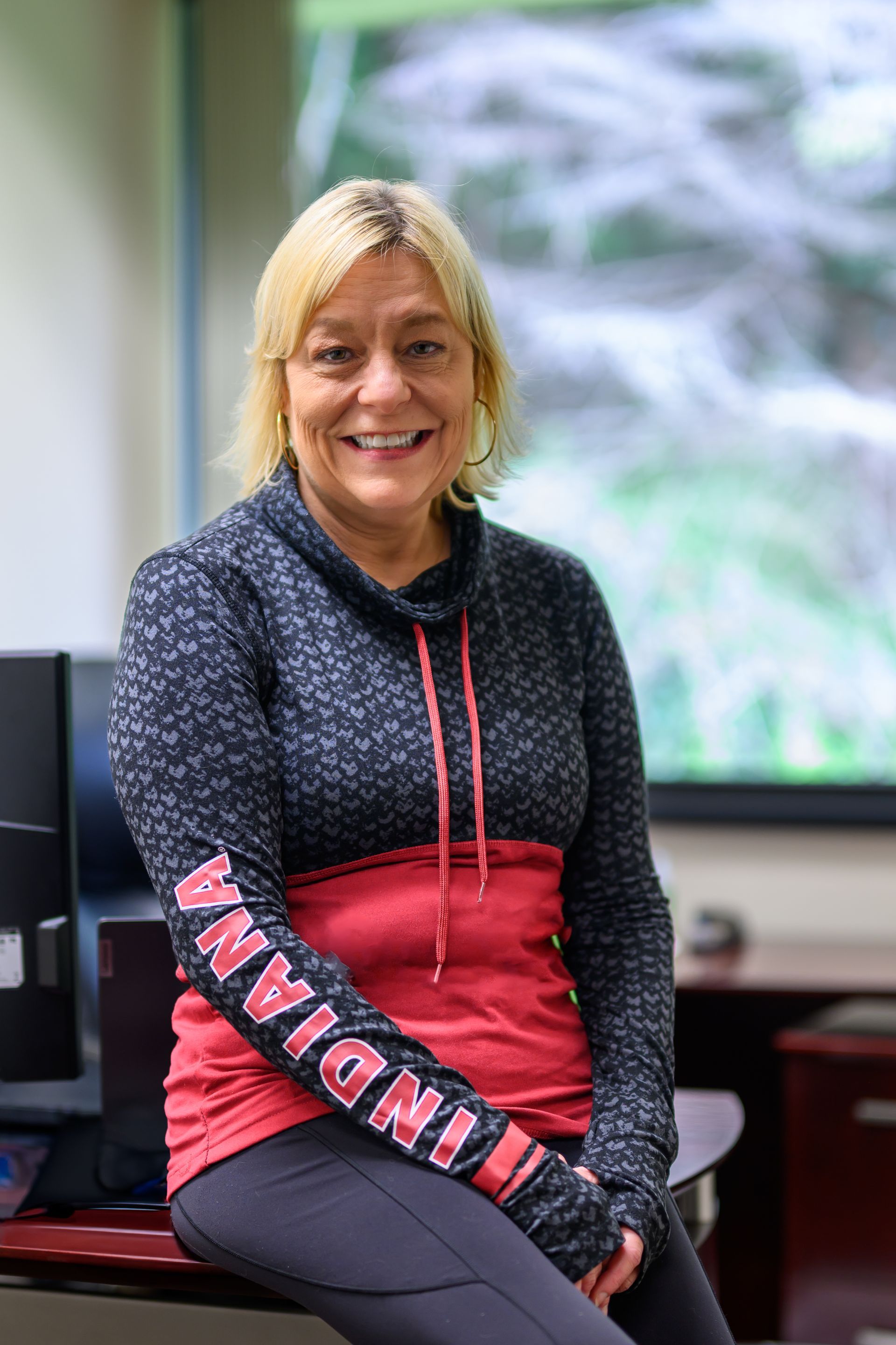 Woman in red and black sweatshirt, sitting on desk, smiling in office.