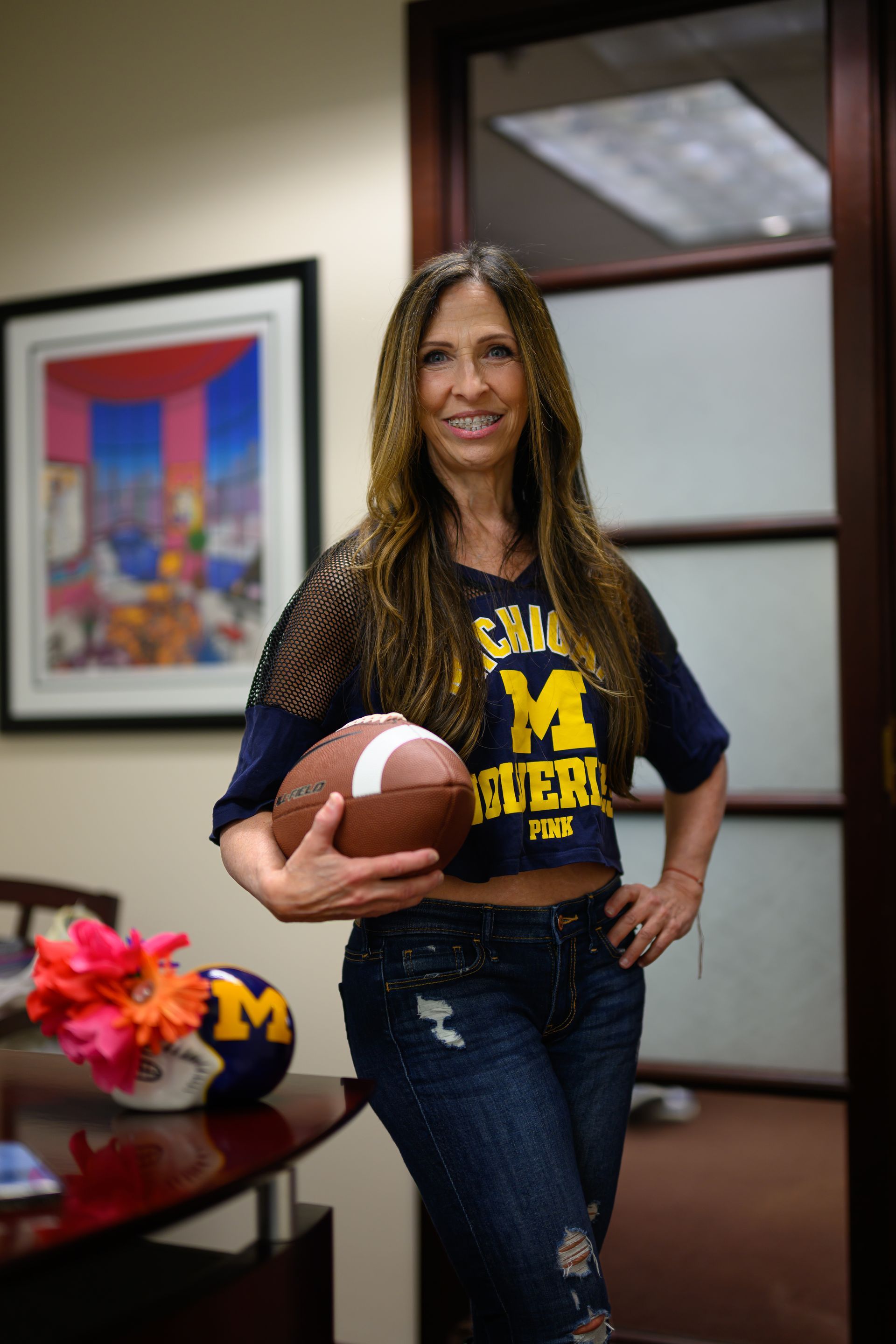Woman holding a football, wearing a Michigan Wolverines crop top and jeans, standing in an office.