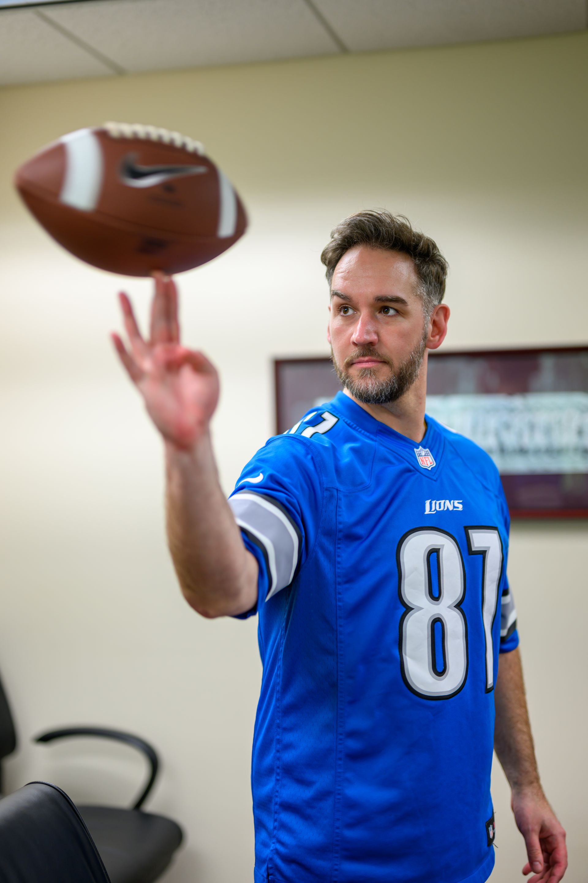 Man in a Detroit Lions jersey balances a football on his finger in an office setting.