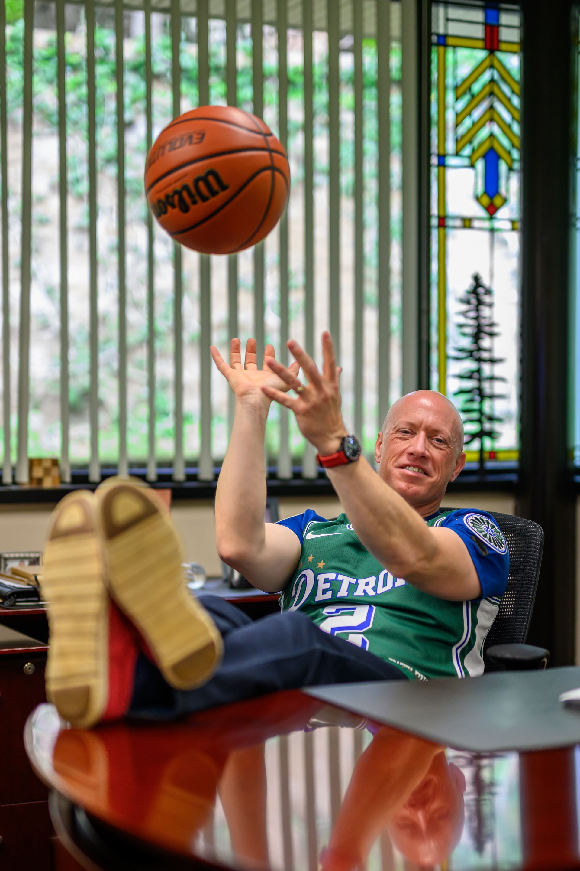 Man tossing a basketball in an office, feet up on a desk. Green shirt, stained glass window background.