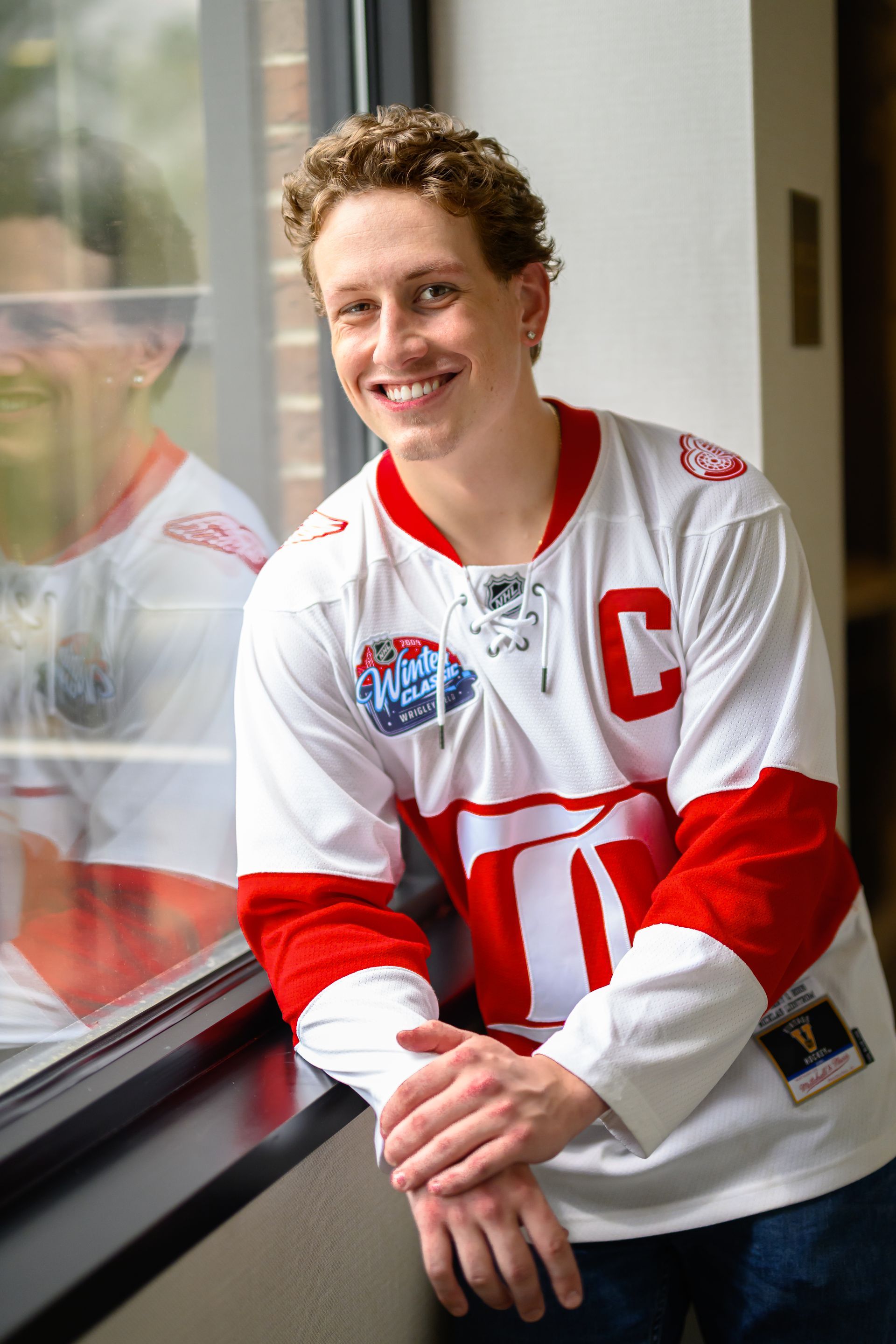 Man in white hockey jersey with red accents leaning against a window, smiling.